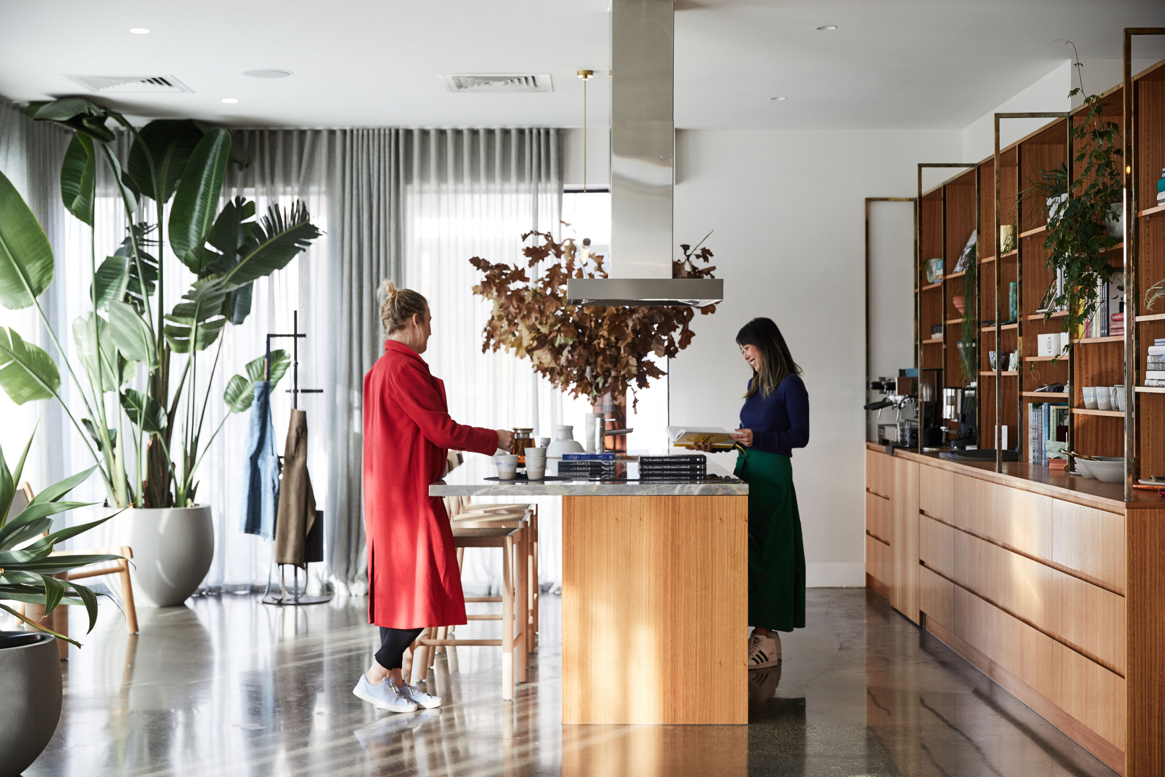 Two women in a stylish kitchen with plants and wooden shelves, chatting at an island.